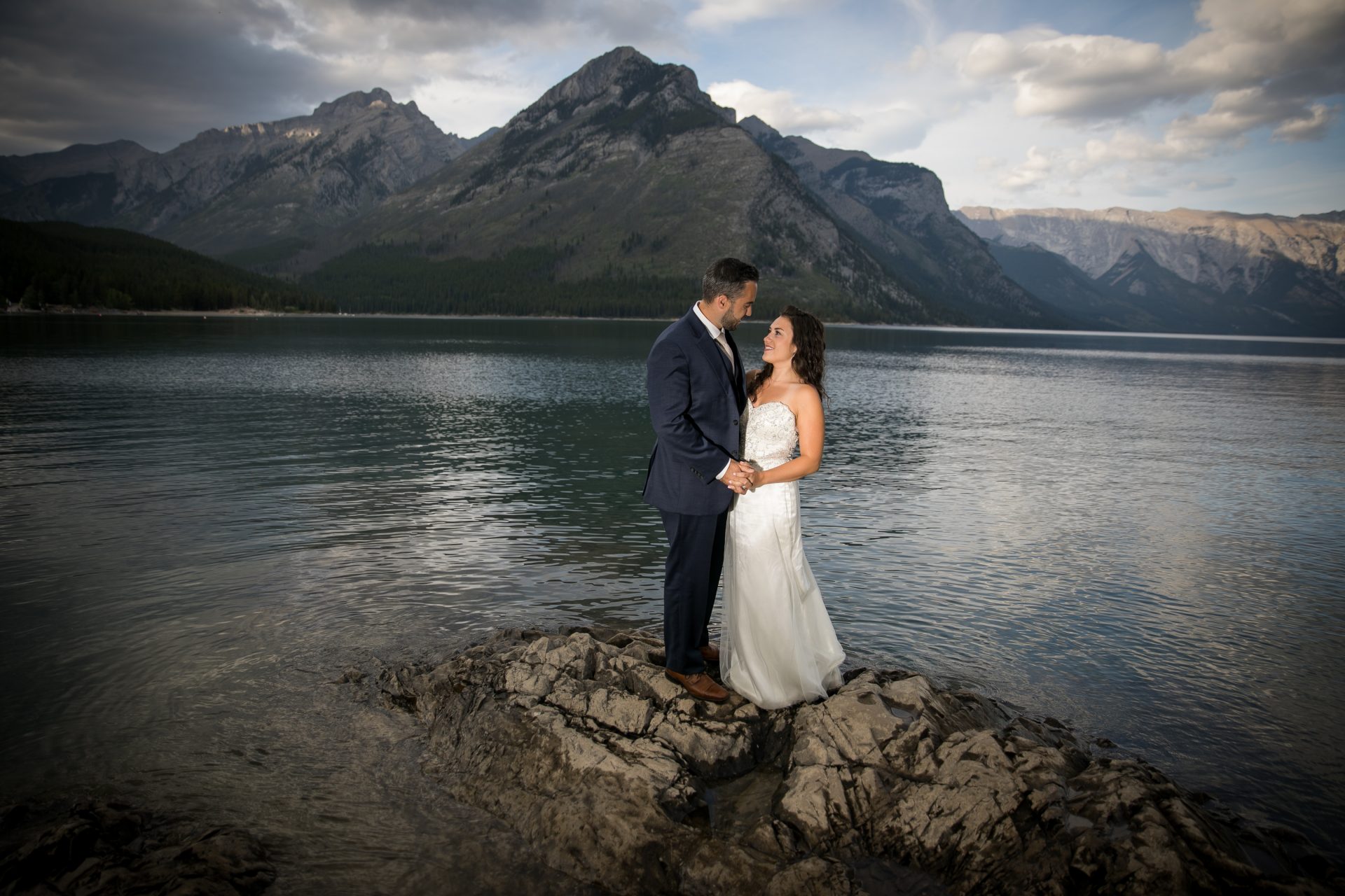 Elegant wedding couple on rocky lakeshore with mountain backdrop in Canmore, Banff, Alberta, captured by Chinook Photography, Expert wedding photographers in Calgary and the Canadian Rockies.