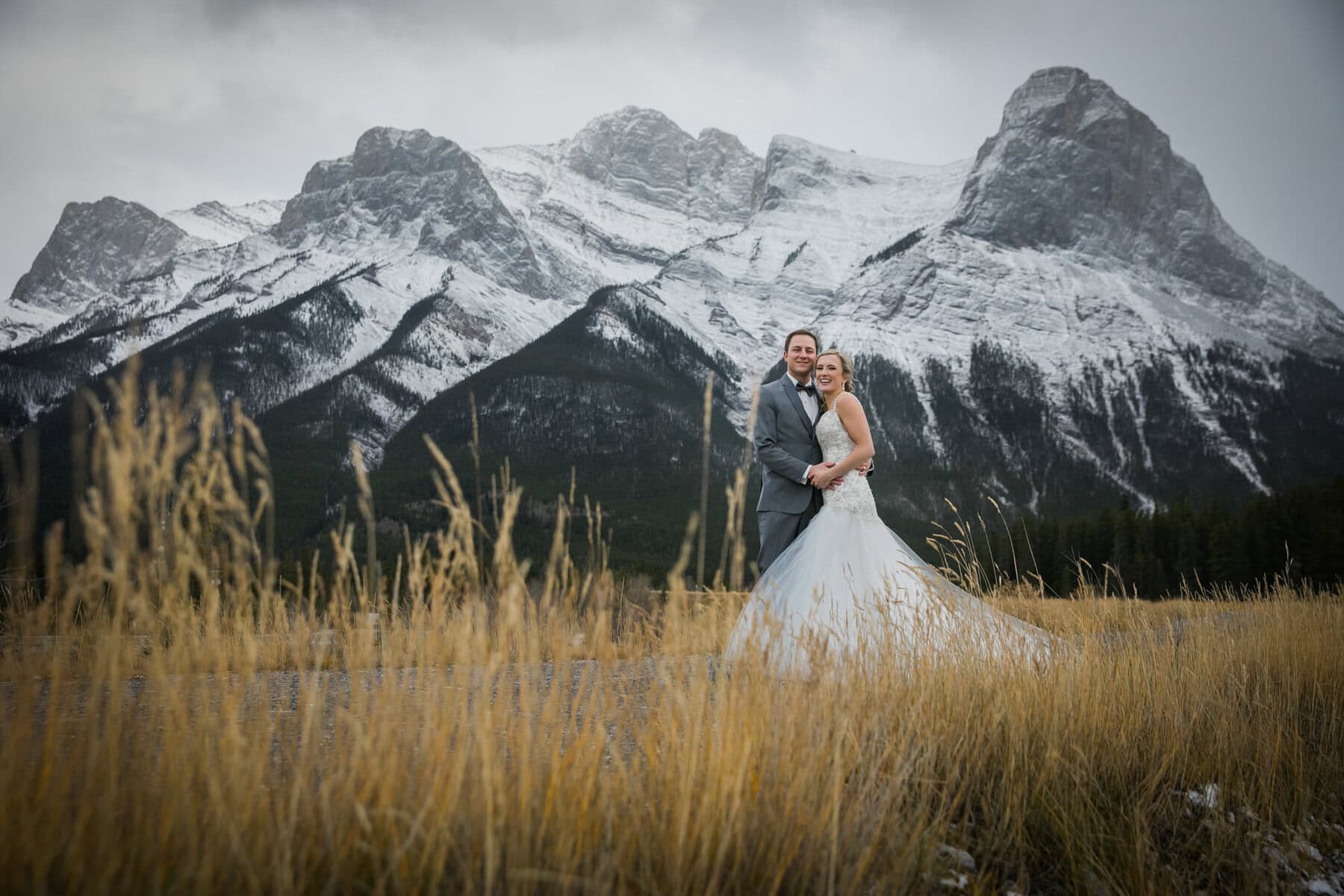 Snow-capped mountains behind a happy wedding couple in Canmore, Alberta, during an outdoor wedding photoshoot with Chinook Photography, featuring Banff wedding photography and scenic Alberta mountain landscapes.